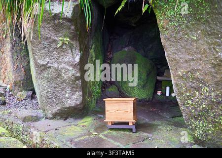 Une boîte d'offrandes en bois traditionnelle positionnée à l'entrée d'un sanctuaire de grotte en pierre couvert de mousse. La scène présente de gros rochers érodés encadrant Banque D'Images