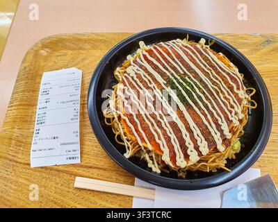 Une vue de dessus d'un okonomiyaki de style Hiroshima ou d'un plat similaire de pancake japonais salé servi sur une assiette en fer noir, posée sur un plateau en bois clair. Banque D'Images