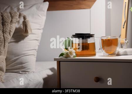 Thé au jasmin dans une théière en verre et une tasse avec des fleurs de jasmin fraîches sur une table de chevet à côté d'un lit, un oreiller blanc et une couverture texturée sont visibles o Banque D'Images