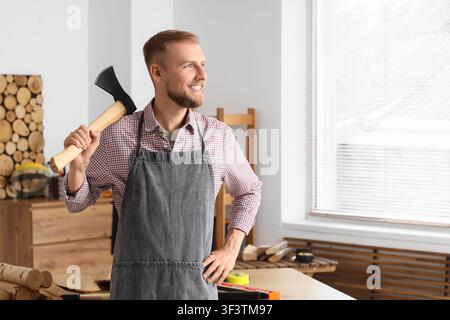 Jeune charpentier souriant avec une hache sur l'épaule dans un atelier de menuiserie, regardant au loin avec une expression heureuse, il porte une chemise à carreaux et une grise Banque D'Images