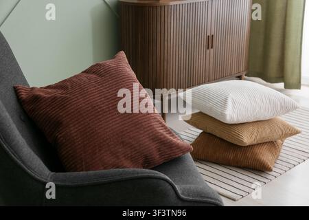 Coussins en velours côtelé dans des tons terreux variés sur un fauteuil gris et un tapis rayé dans un salon moderne, une armoire en bois et un mur vert clair offrent une Banque D'Images