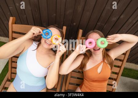Vue aérienne de deux amies joyeuses en maillots de bain allongées sur des chaises longues en bois, souriantes et tenant de petits anneaux gonflables comme des lunettes sur leurs yeux Banque D'Images