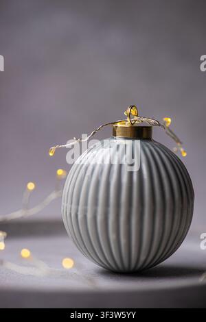 Gros plan d'une boule de Noël rayée grise avec un capuchon doré, enveloppée dans une lumière chaude de guirlande sur un fond gris doux. Nature morte de fêtes minimaliste avec une ambiance douillette Banque D'Images