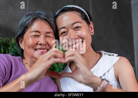 Portrait en gros plan d'une mère souriante et de sa fille adulte posant ensemble et formant un cœur avec leurs mains, montrant l'amour, le lien familial et le bonheur Banque D'Images