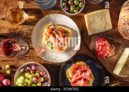 Vue aérienne de tapas méditerranéennes rustiques avec du vin, du pain, du fromage, de la viande séchée, des raisins et des olives sur une table en bois, avec de petites toasts garnies w Banque D'Images