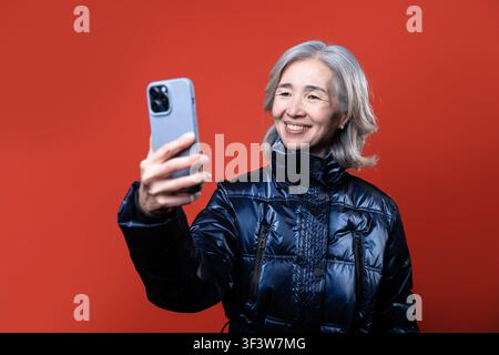 Femme asiatique âgée souriante en veste d'hiver chaude prenant un selfie avec un smartphone devant un fond studio orange, montrant la communication moderne, style de vie Banque D'Images