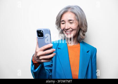 Femme asiatique âgée souriante en blazer bleu tenant un smartphone pour un selfie, profitant d'un appel vidéo ou des réseaux sociaux à la maison devant un fond propre et clair Banque D'Images