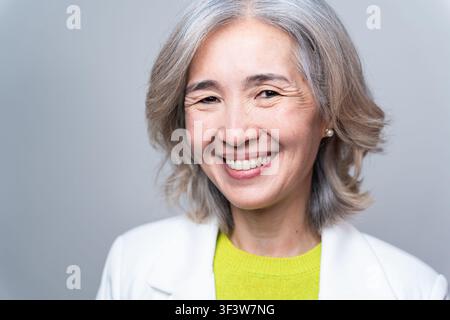 Portrait studio chaleureux d'une femme asiatique âgée souriante aux cheveux gris, portant un blazer blanc et un haut vert, regardant confiante et amicale contre un si Banque D'Images