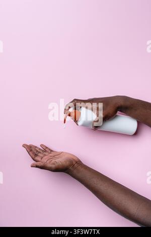 Gros plan des mains d'un homme noir distribuant du savon liquide d'une bouteille à pompe dans la paume ouverte sur un fond rose, mettant en avant l'hygiène, la routine de soins de la peau, un Banque D'Images