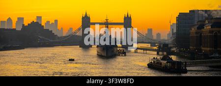 La lumière matinale éclaire la Tamise. Le HMS Belfast est ancré sur l'eau tandis que le Tower Bridge se dresse en silhouette. La ligne d'horizon de la ville dessine la scène, créant une vue visuelle dynamique. Banque D'Images
