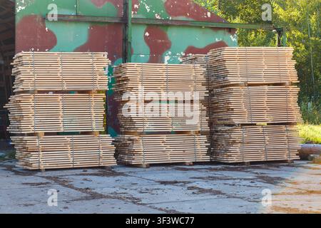 Des piles soignées de planches de bois emballées stockées à l'extérieur sur un sol en béton devant un vieux hangar industriel camouflé dans une scierie. Banque D'Images