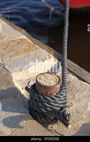 Corde noire solidement attachée autour d'une borne métallique rouillée sur une jetée en béton, offrant un amarrage sûr pour un bateau dans le port avec une eau calme dans le bac Banque D'Images