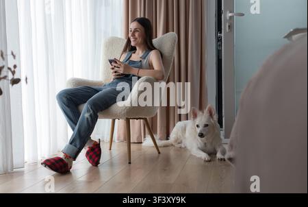 Jeune femme souriante tenant un smartphone tout en se relaxant dans un fauteuil à la maison avec un chien blanc couché à côté d'elle. Banque D'Images
