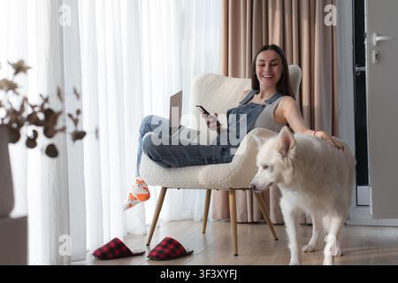 Jeune femme souriante caresse un chien blanc qui passe devant elle tout en tenant un smartphone avec un ordinateur portable ouvert sur ses genoux à la maison. Banque D'Images