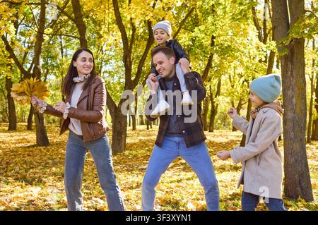 Famille heureuse avec deux enfants jouant parmi les feuilles jaunes d'automne dans un parc ensoleillé Banque D'Images