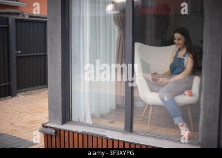 Vue de l'extérieur à travers une grande fenêtre d'une jeune femme souriante travaillant sur un ordinateur portable alors qu'elle est assise dans un fauteuil à l'intérieur d'une maison moderne. Banque D'Images