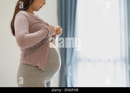 Close-up d'une femme enceinte montrant son ventre de grossesse tout en se tenant près d'une fenêtre. Banque D'Images