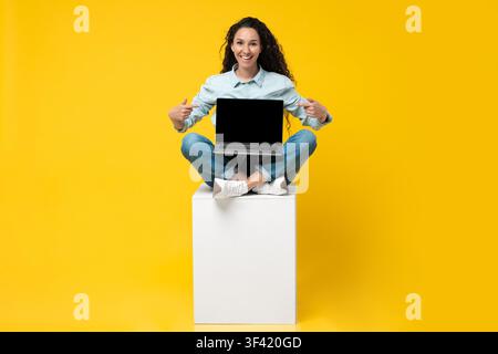 Jeune femme heureuse assise en tailleur sur une boîte, tenant un ordinateur portable avec écran vierge, fond jaune Banque D'Images