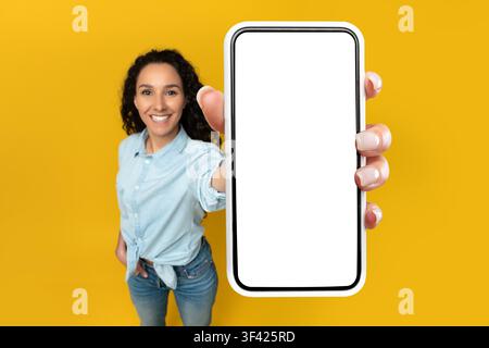 Jeune femme noire souriante tenant un smartphone avec écran vide, fond jaune Banque D'Images