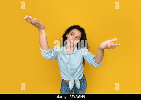 Jeune femme pensive aux cheveux bouclés montrant un fond jaune, se sentant confuse Banque D'Images