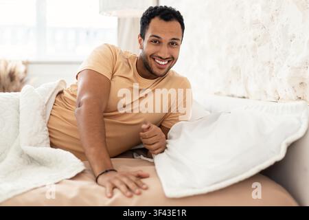 Jeune homme souriant se détendant au lit à la maison, ambiance matinale, concept de mode de vie sain Banque D'Images