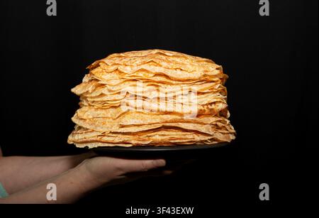 Close-up de mains tenant un énorme tas de crêpes traditionnelles faites maison sur une assiette contre un fond noir foncé. Banque D'Images