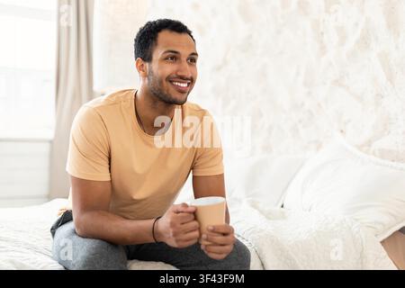 Homme noir souriant assis sur un lit tenant une tasse de café, se détendant à la maison Banque D'Images