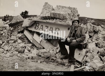 Juste derrière la plage de l'invasion normande, un bazar français est mis hors service, victime des bombardements navals intenses qui ont accompagné l'assaut sur la côte française. Le garde-côte Richard Roig, du 2126 45th Street, Long Island City, New York, fait une pause pour reprendre son souffle au milieu des décombres. L'enseigne brisée du bâtiment, portant l'inscription „Bazar“, gît parmi les maçonneries détruites et les débris éparpillés sur le site. Roig est assis sur les décombres, prenant un moment de calme pendant les conséquences de l'invasion, entouré des ruines de ce qui était autrefois une boutique locale. Banque D'Images