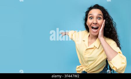Jeune femme excitée aux cheveux bouclés exprimant sa joie devant un fond bleu Banque D'Images