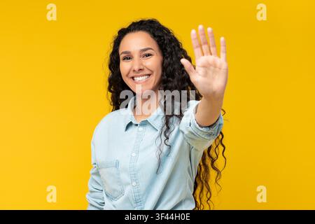 Jeune femme latino-américaine souriante faisant un signe de la main à l'appareil photo, fond jaune Banque D'Images