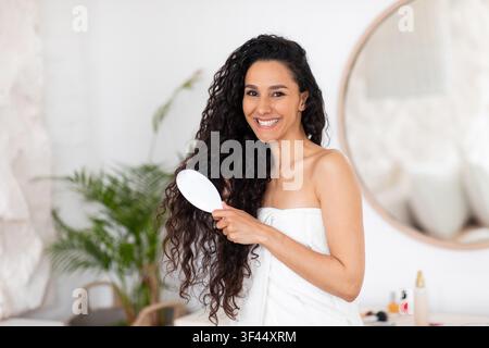 Jeune femme heureuse brossant ses longs cheveux bouclés à la maison, concept de soins de la peau Banque D'Images