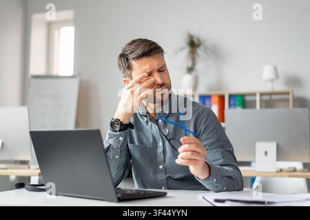 Jeune homme stressé tenant des lunettes et se frottant le front pendant qu'il travaille au bureau, concept d'affaires Banque D'Images