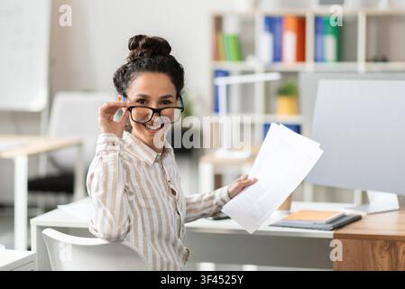 Jeune femme latino-américaine souriante portant des lunettes au bureau, tenant des papiers. Concept d'affaires Banque D'Images