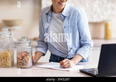 Jeune femme souriante écrivant dans un carnet tout en se tenant dans une cuisine moderne, concept de bureau à domicile Banque D'Images