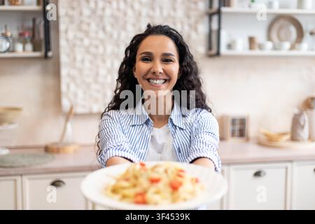 Femme noire souriante tenant une assiette de pâtes dans la cuisine, concept de cuisine joyeuse Banque D'Images
