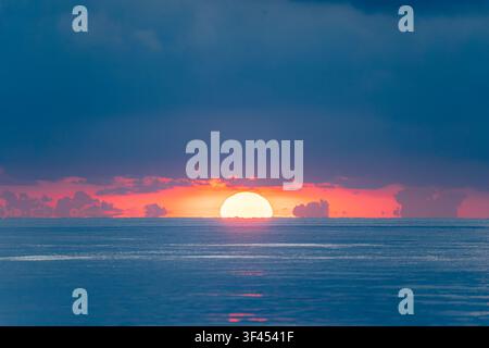 Une vue époustouflante du soleil se couchant sur l'horizon calme de l'océan, projetant une lueur radieuse à travers l'eau et illuminant les nuages dramatiques d'un rouge ardent Banque D'Images