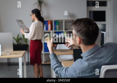 Homme d'affaires prenant une photo d'un collègue avec un smartphone dans un bureau moderne, femme non identifiable Banque D'Images