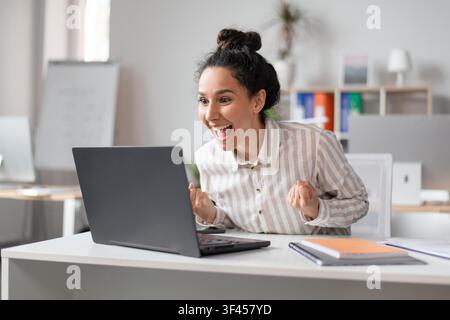 Jeune femme excitée célébrant le succès sur un ordinateur portable dans un bureau moderne, concept commercial Banque D'Images