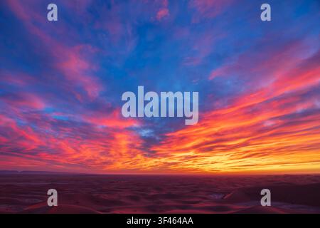 Le ciel au lever du soleil au-dessus des dunes de sable de Chaggaga, de légers nuages reflétant une lumière rouge et orange contre un ciel bleu. Banque D'Images