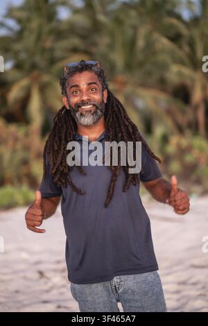 Homme heureux avec des dreadlocks donnant un pouce levé sur une plage Banque D'Images
