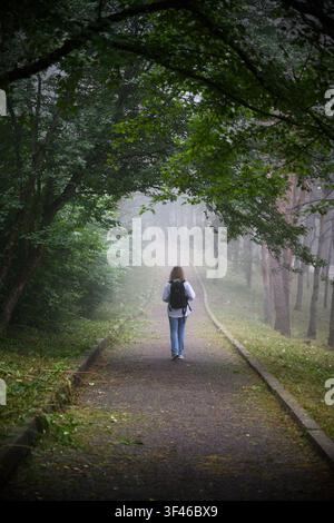 Une photo verticale montrant une femme s'éloignant de l'appareil photo le long d'un chemin pavé à travers une forêt dense. La femme porte une robe de couleur claire Banque D'Images