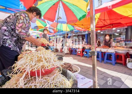 Des pousses de mungo fraîches sont disposées dans des paniers rouges sur un marché asiatique animé. Un vendeur local prépare ces ingrédients essentiels sous des parasols colorés. Banque D'Images