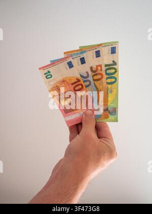 Gros plan d'une main tenant un éventail de billets en euros variés comprenant des billets de 10, 20, 50 et 100 euros sur un fond neutre clair. L'image Banque D'Images