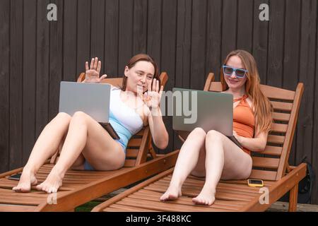 Deux femmes se détendent sur des chaises longues en bois dans un jardin, travaillant sur des ordinateurs portables et bavardant. Travail à distance décontracté en été, temps libre et style de vie en plein air Banque D'Images