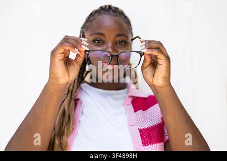 Portrait en gros plan d'une femme noire aux longues tresses soulevant ses lunettes et regardant l'appareil photo, portant un cardigan rose et un haut blanc contre un brig Banque D'Images