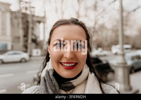 Portrait souriant d'une femme heureuse avec du rouge à lèvres rouge et une écharpe à carreaux, posant en plein air sur une rue urbaine floue Banque D'Images