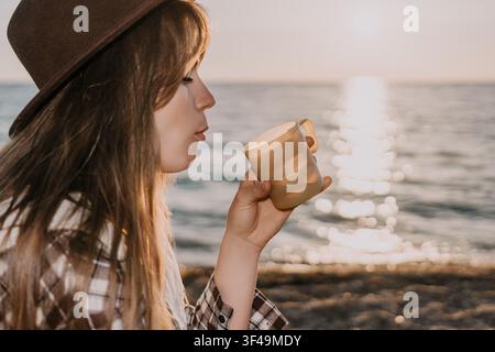 Femme café plage profitant d'une boisson chaude dans une tasse au coucher du soleil tout en étant assise sur la côte océanique en vacances Banque D'Images