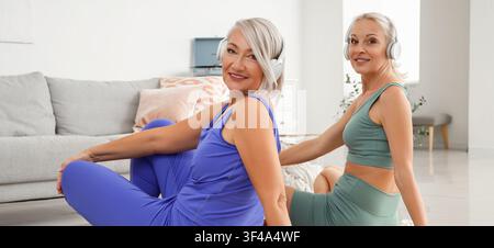 Deux femmes âgées souriantes avec des écouteurs, assises par terre en vêtements de sport, profitant d'un mode de vie sain et de la forme physique à la maison Banque D'Images