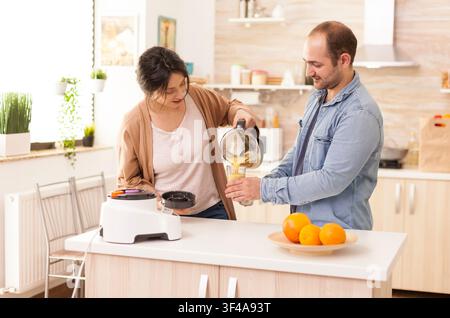 La femme verse un smoothie nutritif dans des verres pour elle et son mari, suit un régime et prépare le petit-déjeuner dans un matin ensoleillé et confortable, reliant le petit-déjeuner en cuisine, la cuisine domestique et la routine matinale. Banque D'Images