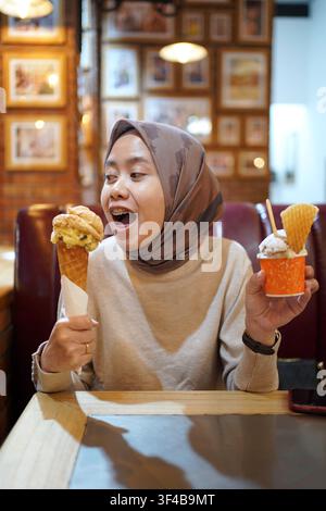 Femme indonésienne en hijab tenant une glace en cornet et une tasse tout en savourant un dessert dans un café. Concept de nourriture amusante et de style de vie. Banque D'Images